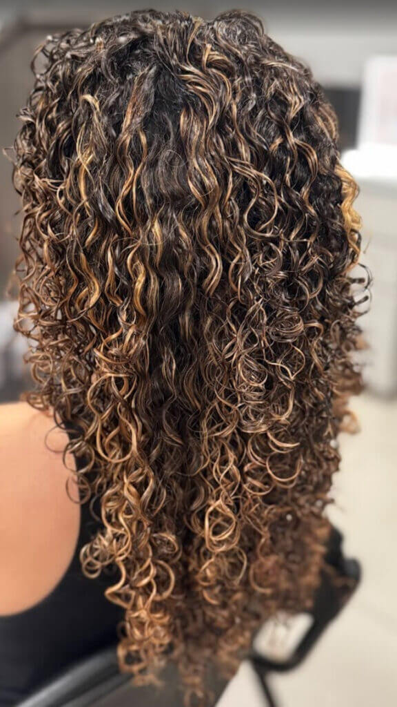 Cabelo cacheado com mechas moreno iluminado, efeito natural e brilho intenso, realizado no Studio Jessica Miranda, salão de beleza em Cabo Frio.