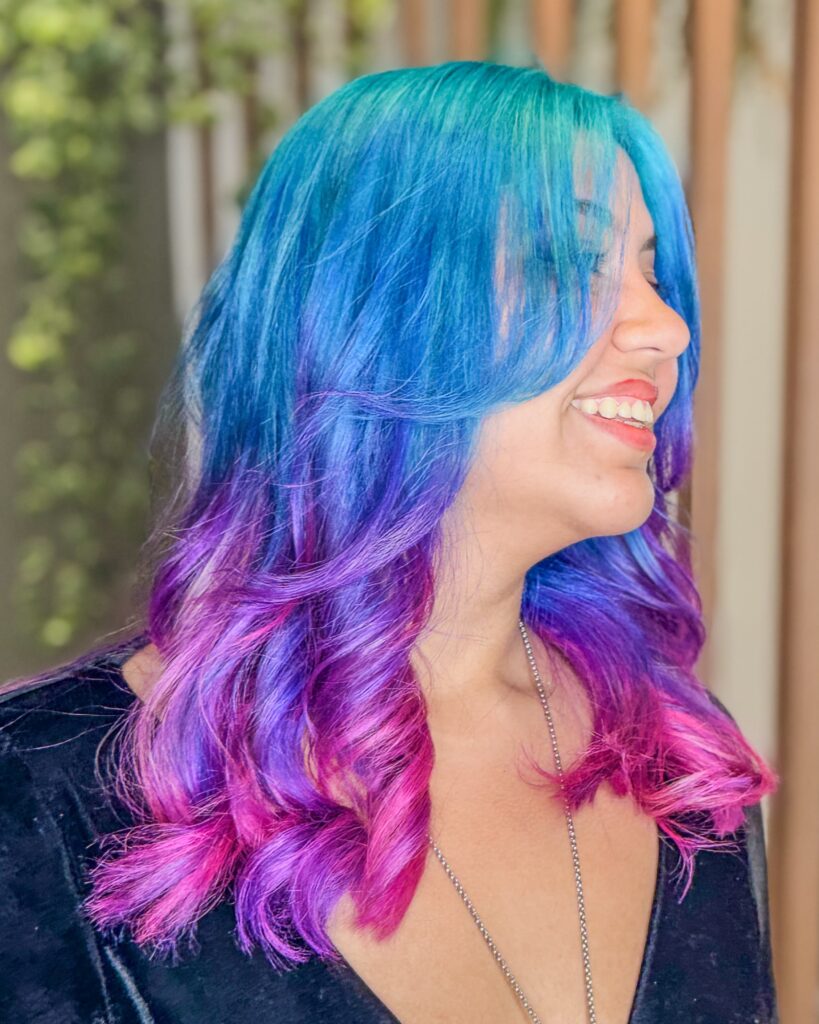 Coloração fantasia em estilo glitch hair, com cabelo azul vibrante na parte superior e nuances de roxo e pink no comprimento e nas pontas, realizada no Studio Jessica Miranda, salão de beleza em Cabo Frio.