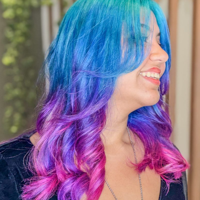 Coloração fantasia em estilo glitch hair, com cabelo azul vibrante na parte superior e nuances de roxo e pink no comprimento e nas pontas, realizada no Studio Jessica Miranda, salão de beleza em Cabo Frio.